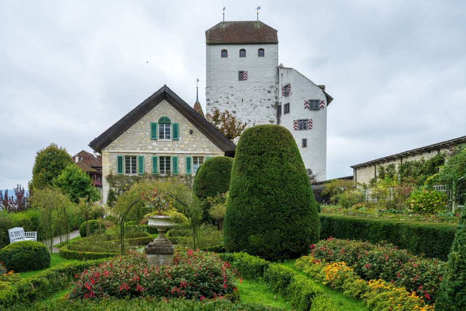 A well-maintained garden featuring neatly trimmed hedges and topiary bushes in front of a historic white castle-like building with small windows and a tower, set against a cloudy sky. The garden beds are filled with colorful flowering plants and are bordered by low, manicured grass edges. A paved pathway runs through the lush greenery, with a classical stone birdbath or planter situated in the center of the flower beds. Mature trees with dense foliage surround the area, adding to the garden's lush appearance. Visible tools, such as a garden rake and shears, indicate ongoing lawn care and hedge trimming work. The scene demonstrates careful landscaping and outdoor maintenance, typical of garden care services offered by Gardeners Swiss Cottage, emphasizing their expertise in garden maintenance and landscaping work.