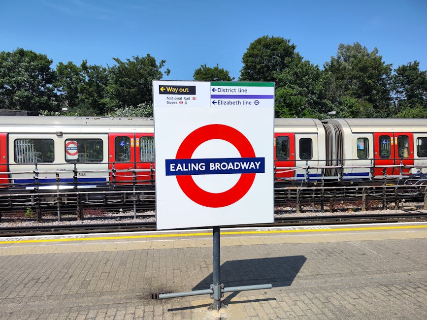 A London Underground informational sign in a garden setting at Ealing Broadway station, with a train parked on the tracks behind it. The sign displays station details, including directions to the way out, district line, Elizabeth line, and nearby bus connections, and is mounted on a metal stand on a paved platform. In the background, there are green trees and a clear blue sky, providing a natural environment contrast to the urban transit scene. The surroundings suggest a well-maintained outdoor area associated with the station, possibly featuring landscaped greenery, and this image exemplifies outdoor maintenance of transport hubs, aligning with gardening and landscaping themes that Gardeners Swiss Cottage might oversee.