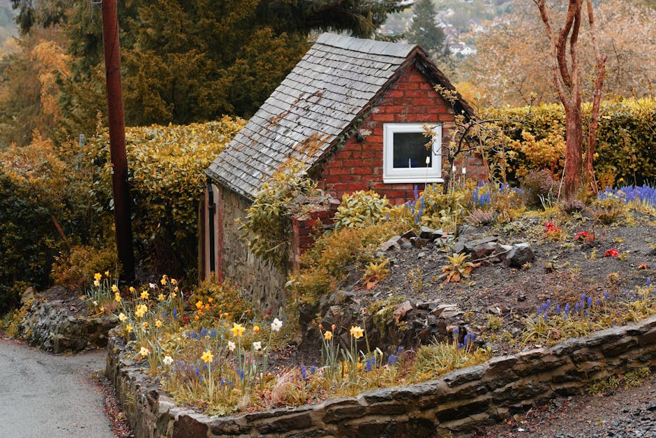 A small garden scene featuring a red brick and stone outbuilding with a pitched tile roof. Surrounding the building are lush, well-maintained shrubs and trees with colorful autumn foliage. The foreground showcases a flower bed edged with a low stone wall, filled with blooming yellow daffodils, purple and blue flowers, and some red blooms, with moist, dark soil visible. To the left, a narrow paved pathway borders the garden, with a wooden utility pole partially visible. The garden is illuminated by soft natural light, highlighting the textures of the plants, soil, and stonework. Gardeners Swiss Cottage is likely tasked with maintaining this garden, emphasizing landscaping work, tidy lawns, and hedge trimming to preserve its neat appearance.